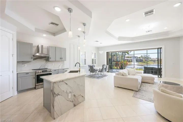 Open-concept living area with large format tile flooring, a kitchen island with stone waterfall edge, stainless steel appliances, and tray ceilings