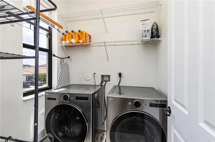 Dedicated laundry room featuring side-by-side front-load washers and dryers with stainless steel finishes, wire shelving with organized storage for cleaning supplies, and large windows providing natural light and ventilation