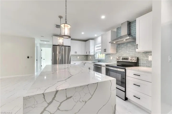 Kitchen with pendant lighting, appliances with stainless steel finishes, white cabinets, light stone counters, and wall chimney range hood