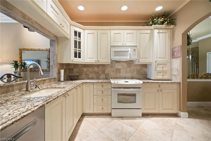 Kitchen featuring cream cabinetry, white appliances, arched walkways, light stone counters, and ornamental molding