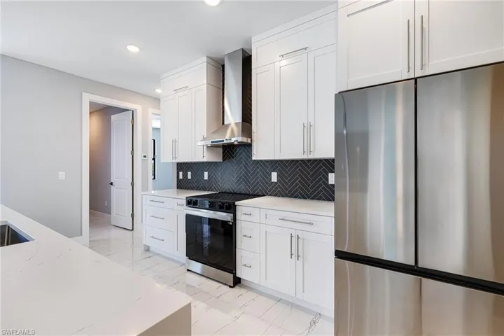 Kitchen featuring stainless steel refrigerator, range with electric stovetop, wall chimney range hood, light marble finish floors, and recessed lighting
