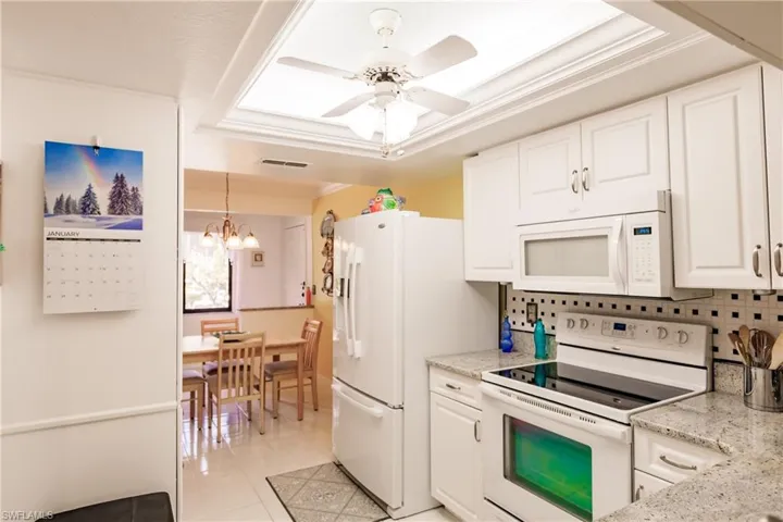 Kitchen with ceiling fan with notable chandelier, white appliances, light stone counters, decorative backsplash, and a raised ceiling