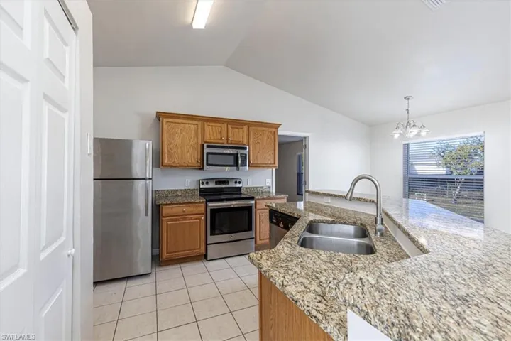 Kitchen with stainless steel appliances, brown cabinets, vaulted ceiling, pendant lighting, and light stone counters