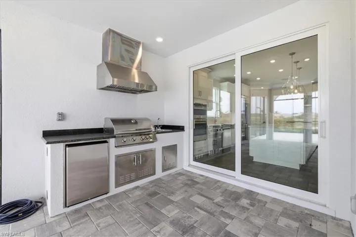 Outdoor kitchen featuring a built-in stainless steel grill, stainless steel range hood, stainless steel undercounter refrigerator, integrated sink, and dark stone countertop