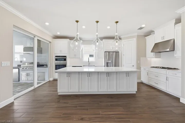 Spacious kitchen featuring an expansive central island with three pendant lights, white cabinetry, integrated stainless steel appliances, and wood-finish flooring