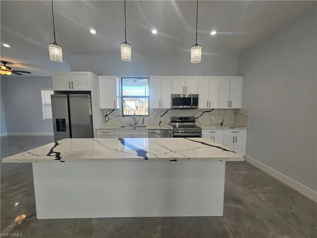 Kitchen with light stone countertops, decorative backsplash, stainless steel appliances, white cabinetry, and pendant lighting
