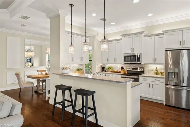 Kitchen featuring a kitchen breakfast bar, light stone counters, custom pendants, stainless steel appliances, dark hardwood floors, and white cabinetry with custom backsplash tile