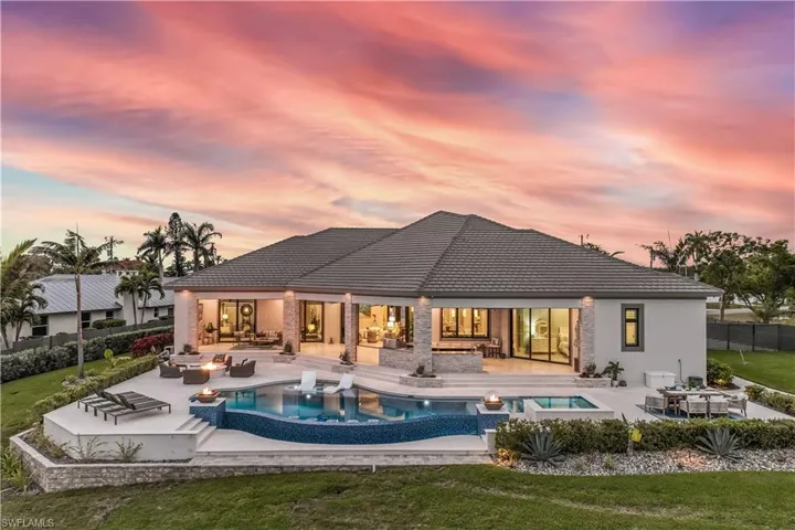Back house at dusk featuring an outdoor living space with a fire pit, exterior kitchen, a swimming pool with hot tub, a patio area, and a yard