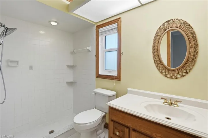 Bathroom featuring a tiled shower, overhead lighting, a window with wood trim, a wood-finish vanity with an integrated sink, and an oval mirror