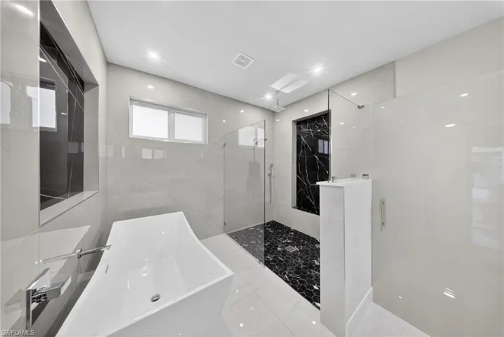 Contemporary bathroom featuring a freestanding soaking tub with a wall-mounted chrome faucet, a glass-enclosed shower with black hexagonal floor tiles, and light-toned tile walls