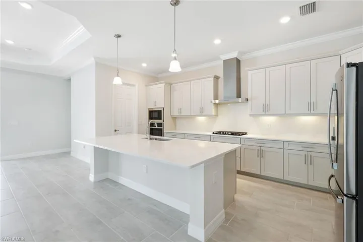 Kitchen featuring hanging light fixtures, stainless steel appliances, a center island with sink, wall chimney range hood, and ornamental molding
