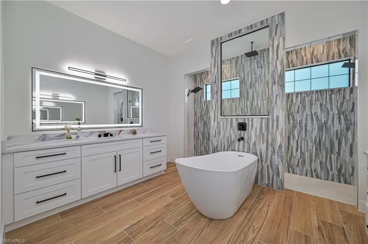 Freestanding soaking tub, wood-finish flooring, dual shower heads, illuminated vanity mirror, and textured wall tile