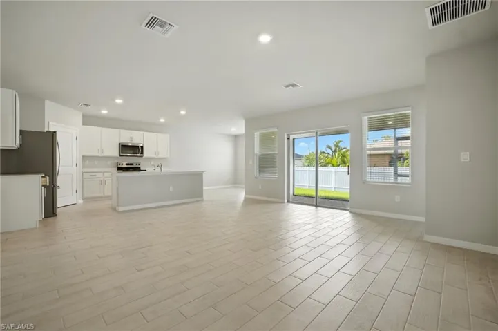 Spacious open-concept living area featuring light-toned tile flooring, recessed lighting, white kitchen cabinetry with stainless steel appliances, a kitchen island with a breakfast bar, and sliding glass doors to the exterior