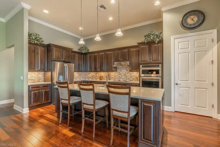 Kitchen with stainless steel appliances, crown molding, dark wood-type flooring, pendant lighting, and an island with sink