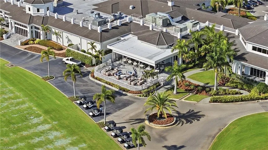 Expansive complex featuring a covered stone patio, tiered shingle roofing, paved golf cart staging area, and manicured grounds with mature palm trees