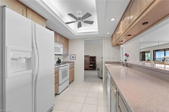 Kitchen with white appliances, a raised ceiling, light stone counters, ceiling fan, and recessed lighting