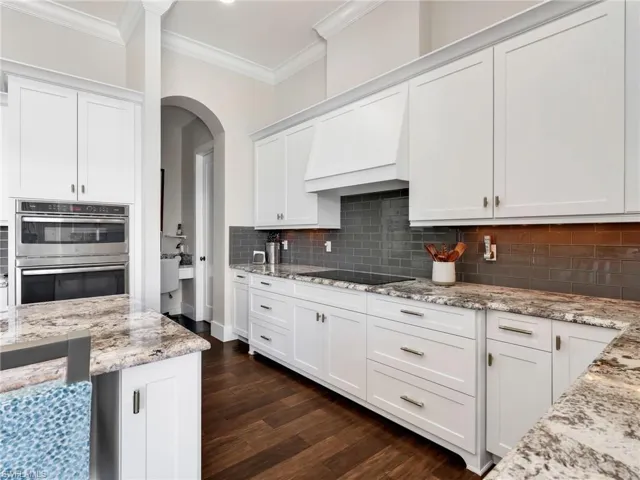 Kitchen with white cabinets, dark wood-style floors, stainless steel double oven, light stone counters, and ornamental molding