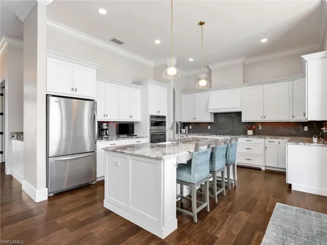 Kitchen with stainless steel appliances, light stone countertops, an island with sink, white cabinetry, and crown molding
