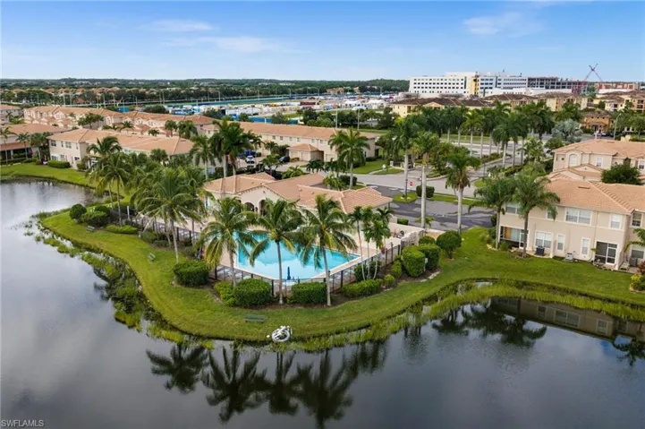 Community swimming pool with surrounding fence, mature palm trees, and residential buildings with barrel tile roofs
