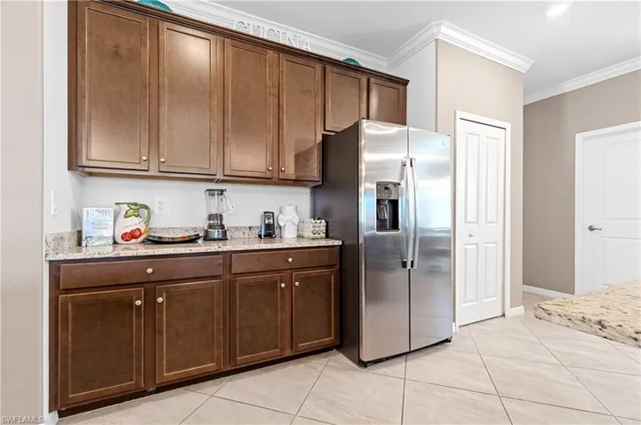 Kitchen featuring stainless steel fridge with ice dispenser, ornamental molding, light stone counters, light tile patterned floors, and dark brown cabinetry