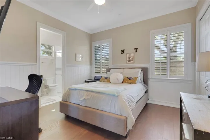 Bedroom featuring wainscoting, wood finished floors, ornamental molding, ceiling fan, and ensuite bath