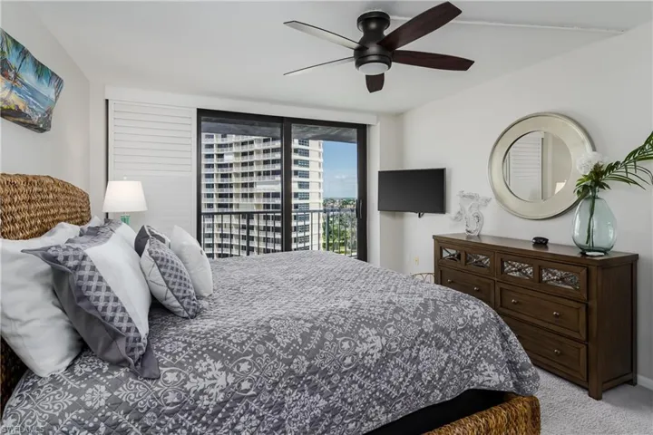 Sliding glass doors with balcony access, ceiling fan with integrated lighting, wall-mounted television, and wood-finish dresser with decorative hardware