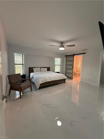 Bedroom featuring a barn door, light marble finish flooring, a ceiling fan, and ensuite bathroom
