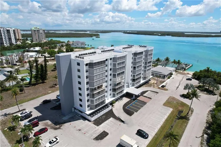 Multi-story waterfront condominium building featuring an expansive parking area and surrounding tropical foliage