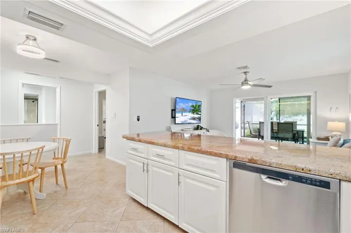 Kitchen with stainless steel dishwasher, light stone countertops, and white cabinets, with open dining area.