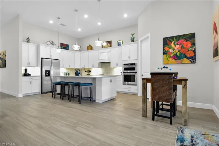 Kitchen featuring appliances with stainless steel finishes, a towering ceiling, white cabinets, hanging light fixtures, and an island with sink