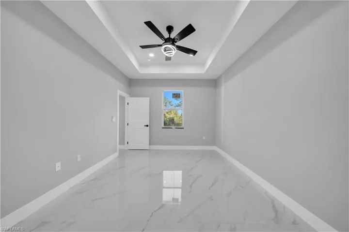 Spacious room featuring a tray ceiling with a modern black ceiling fan, large format marble-look tile flooring, light gray wall paint, and white trim