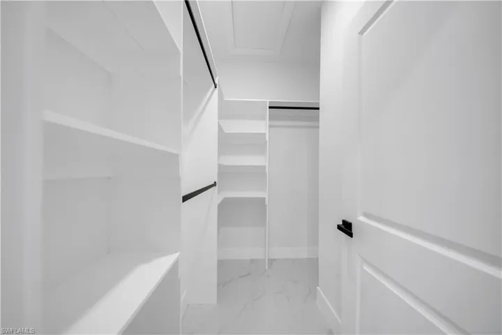 Walk-in closet featuring white built-in shelving, black hanging rods, a white paneled door with black hardware, and large format tile flooring