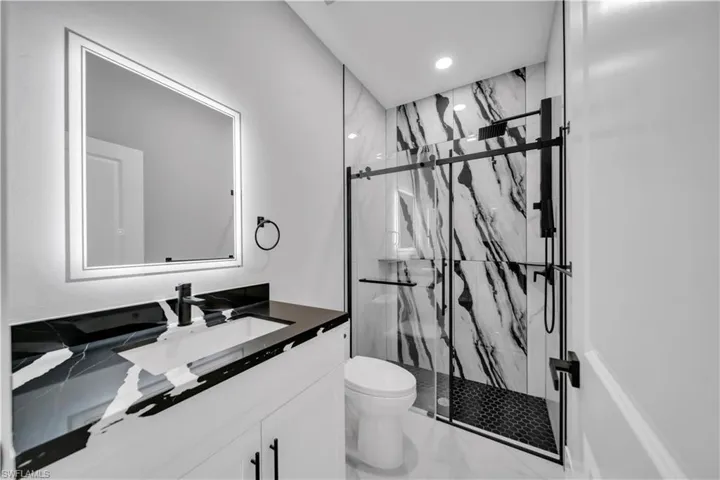 Bathroom featuring a black and white patterned countertop, white vanity with black hardware, back-lit mirror, and a glass-enclosed shower with black fixtures and hexagonal floor tiles
