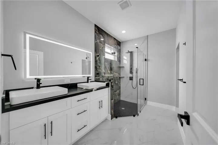 Bathroom featuring dual vessel sinks on a dark countertop, an illuminated mirror, a glass-enclosed shower with dark tile flooring, and white cabinetry with dark hardware