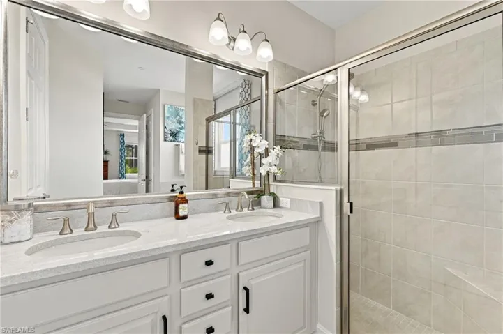 Spacious tiled shower and double vanity featuring white cabinetry, stone-finish countertops, dual undermount sinks, and a full-width mirror with integrated lighting