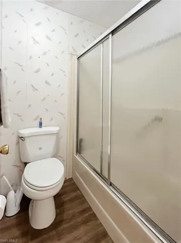 Full bathroom with wallpapered walls, bath / shower combo with glass door, dark wood-style floors, and a textured ceiling