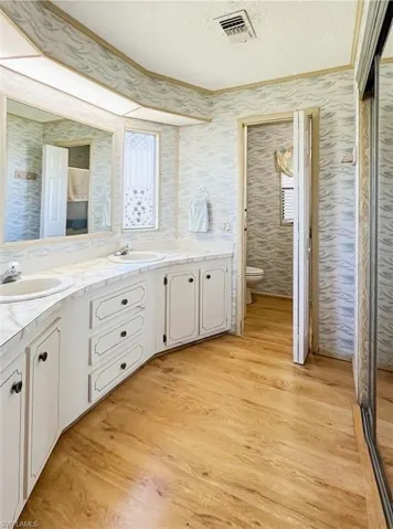 Bathroom with double vanity, a textured ceiling, light wood-style floors, ornamental molding, and wallpapered walls
