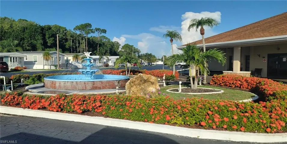 Tiered stone fountain with integrated basin, surrounded by manicured landscape beds featuring mature palm trees and colorful shrubbery