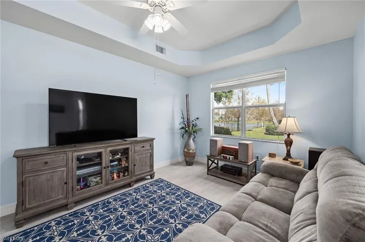 Living area with a raised ceiling, light wood finished floors, baseboards, visible vents, and ceiling fan