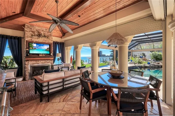 Expansive outdoor living area featuring a stone-clad fireplace with built-in media, wood-finish vaulted ceilings with exposed beams, and a ceiling fan