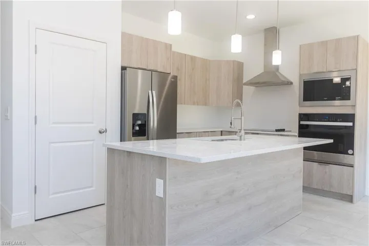 Kitchen featuring light brown cabinets, light stone countertops, modern cabinets, stainless steel appliances, and wall chimney exhaust hood