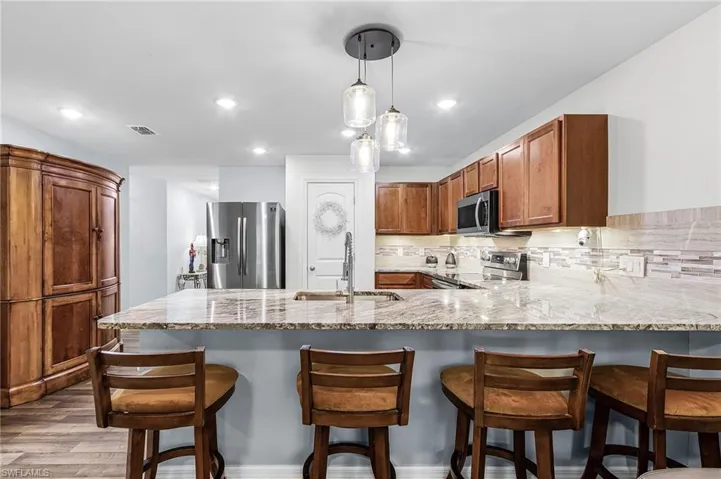 Kitchen with brown cabinetry, decorative backsplash, appliances with stainless steel finishes, light stone counters, and decorative light fixtures