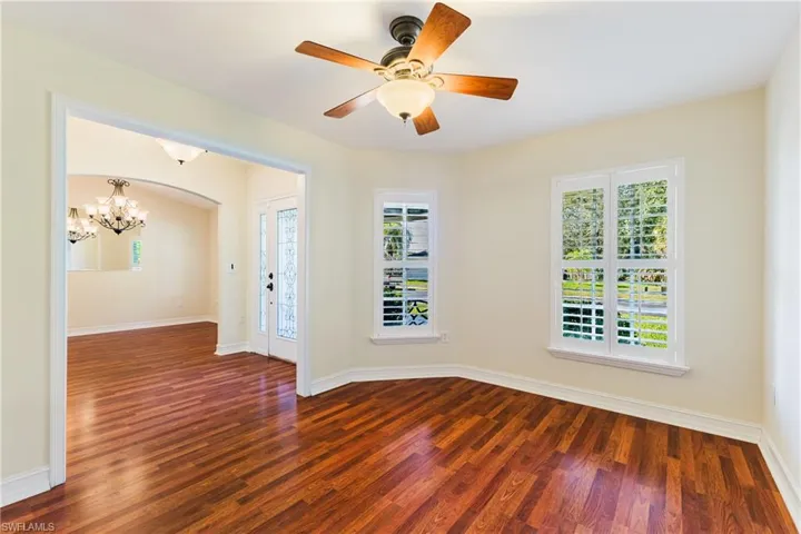 Room featuring wood-finish flooring, two large windows with white louvered shutters, a ceiling fan with light fixture, and an arched doorway leading to an entryway with double glass doors