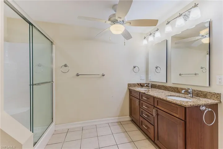 Dual vanity with granite countertop and integrated sinks, glass-enclosed shower, white tile flooring, and ceiling fan with integrated lighting