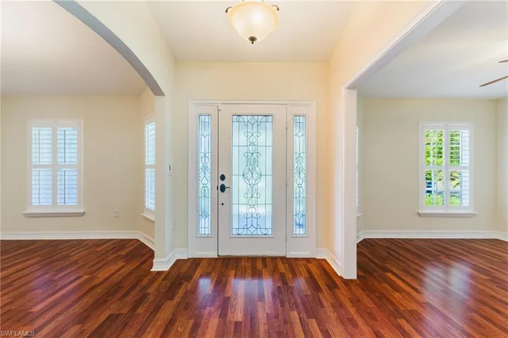 Foyer entry with an ornate glass-paneled door and sidelights, featuring wood-finish flooring, plantation shutters, an arched entryway, and a semi-flush ceiling fixture