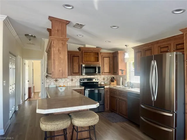 Kitchen featuring stainless steel appliances, a peninsula, brown cabinets, light countertops, and dark wood-style floors