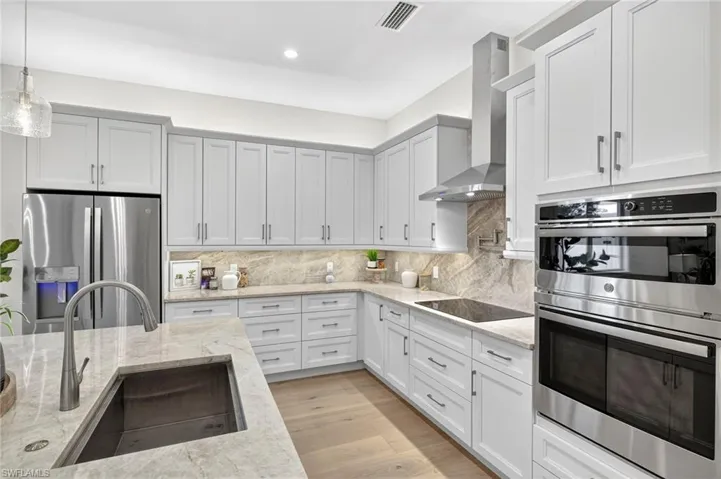 Kitchen with appliances with stainless steel finishes, backsplash, light stone counters, wall chimney range hood, and pendant lighting