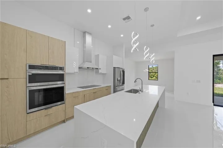 Kitchen with a large island with sink, two tone color scheme, light stone counters, stainless steel appliances, and decorative light fixtures