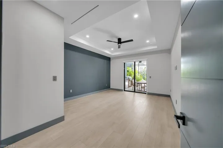 Spacious interior with light wood-finish flooring, a tray ceiling featuring recessed lighting and a ceiling fan, a contrasting accent wall, and sliding glass doors opening to an outdoor patio with a dining area