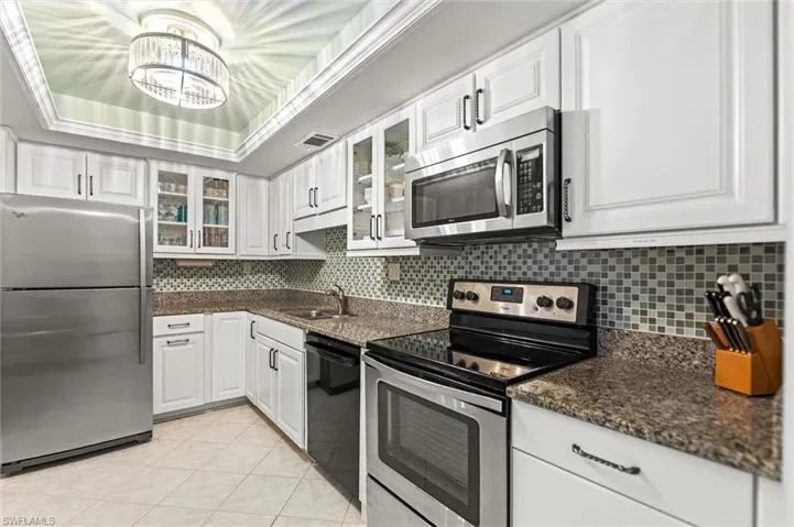 Kitchen with stainless steel appliances, glass fronted cabinets, white cabinetry, dark stone countertops, and a raised ceiling